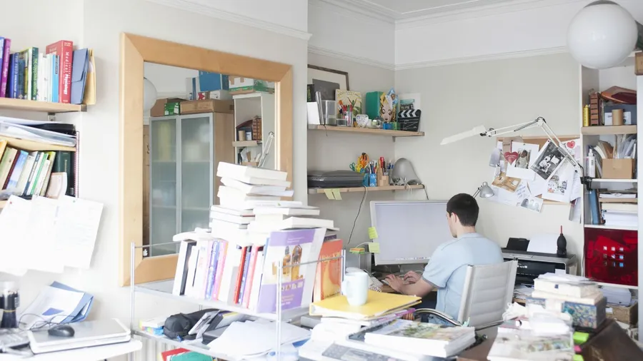 A cluttered room with a mirror on the wall and a man working on a computer.