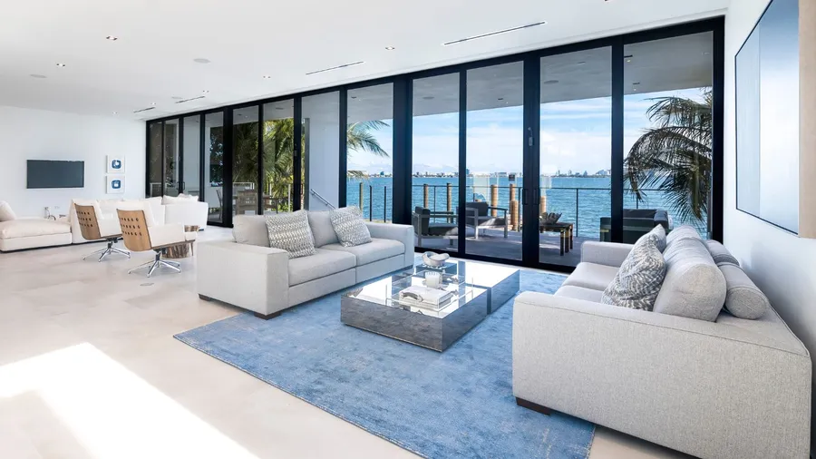A living room interior in coastal color palette with light blue rug, gray sofas with blue and white patterned cushions. and transparent glass doors.