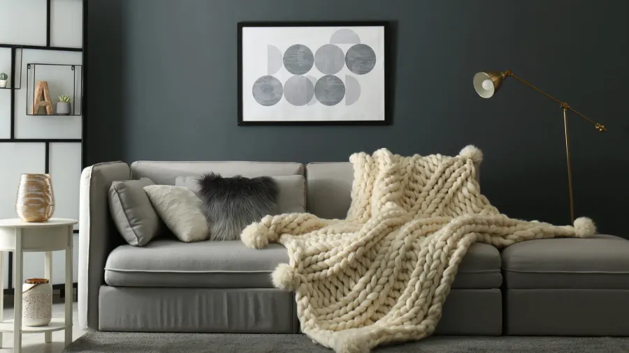 A gray sofa with textured throw pillows and knitted throw blanket, white side table with vases, brass floor lamp, and framed artwork on a gray wall in a living room.