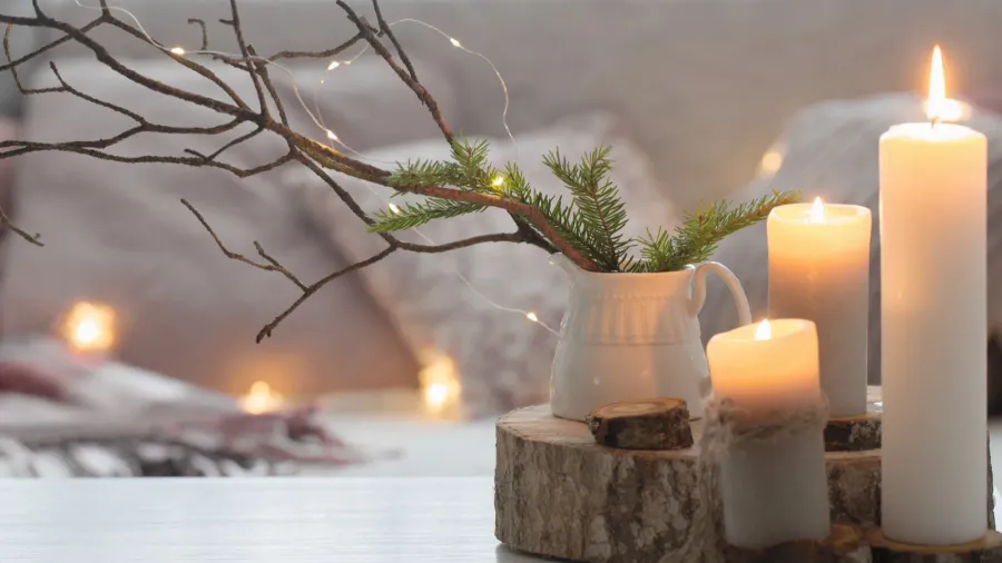 Composition of candles on wooden platters on a while table. A small white vase with plants and branches and fairy lights and sofa in the background.