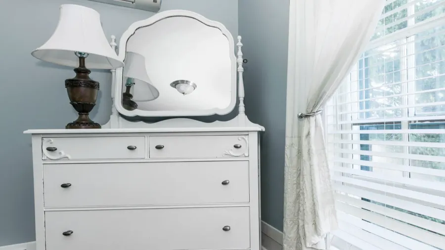 A white dresser with a mirror and lamp on top of it in front of a window.