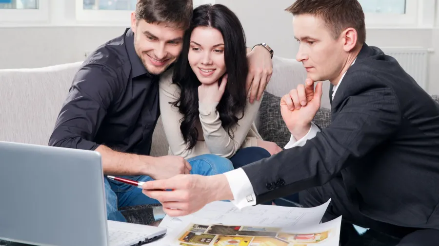 A young couple looking at the laptop as the interior designer explains something to them, pointing with a pen on the screen.