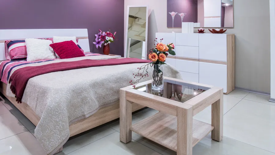 A cozy bedroom featuring a bed with decorative pillows, a stylish bedspread, and a modern nightstand with a glass top. There's a vibrant flower arrangement next to the bed and a white dresser in the background, against a soft purple wall.