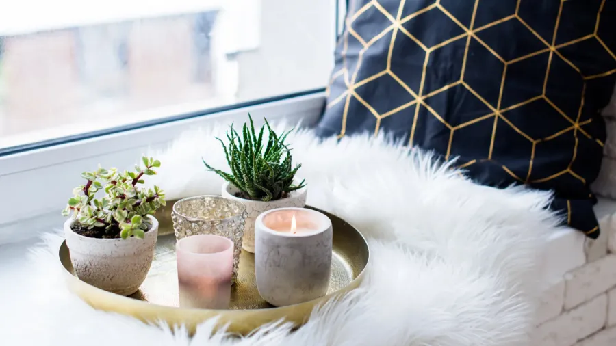 A decorative golden tray with candles and planters on a faux fur surface, and a throw pillow placed on a windowsill.