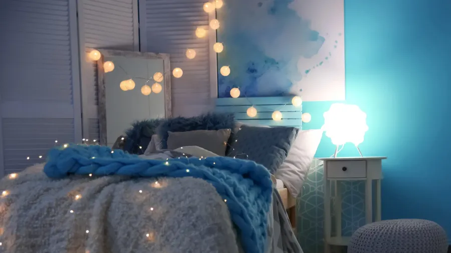 A cozy bedroom interior with comfortable bedding, fairy light canopy, standing mirror, and a small side table with a lamp.