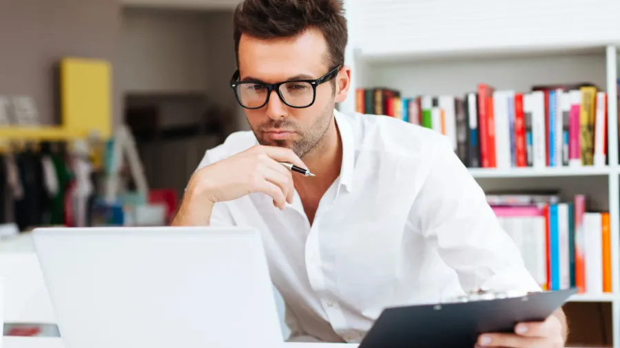 An interior designer holding a pen and a board in his hand, and looking at the screen with concentration.