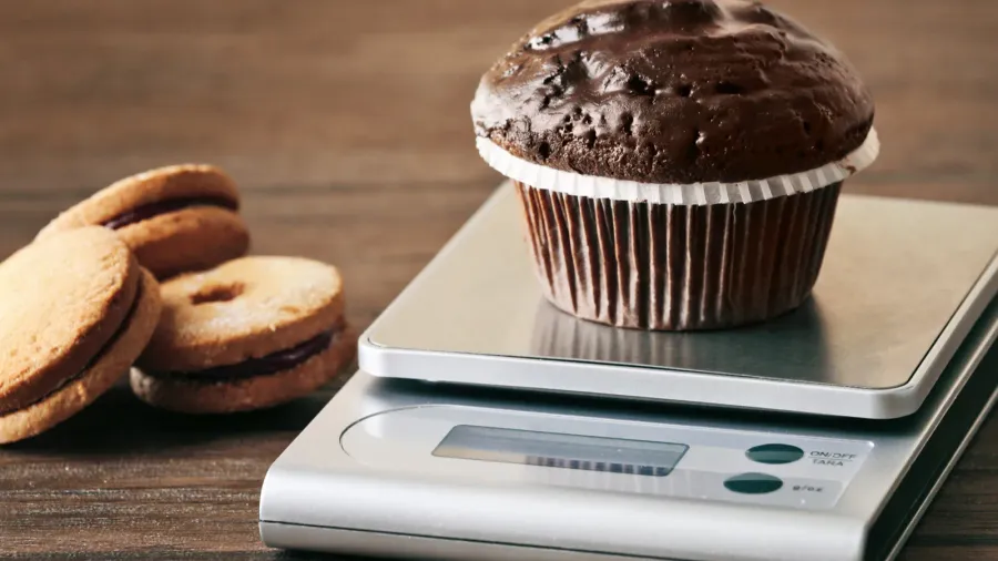 A chocolate muffin on a digital kitchen scale with sandwich cookies on the side.
