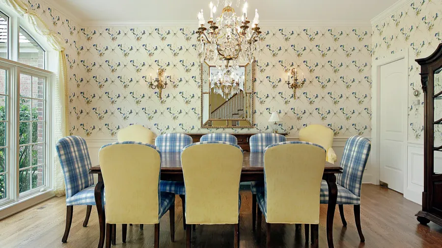 A dining room interior with checkered chairs, wooden table, chandelier, and wallpaper on the walls.