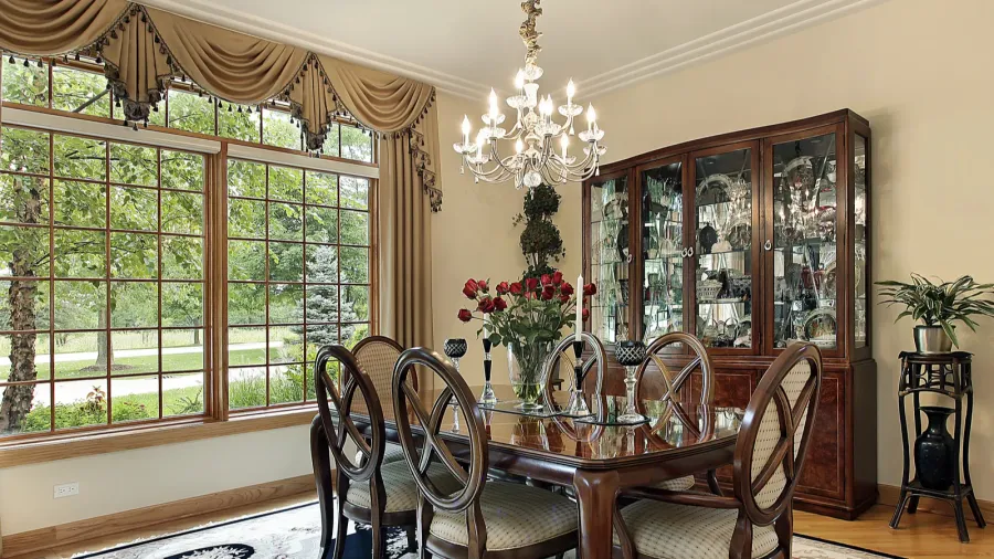 An elegant beige dining room interior with a dark wood furniture, patterned chairs, curtains, shelf displaying dinnerware, and pendant lighting.