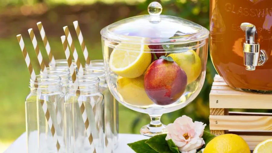 A drink station with glass jars with cute paper straws, fresh fruits, flowers, and a cooler with drink.