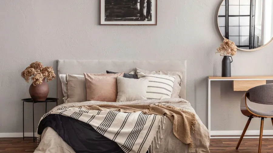 Bedroom interior in neutral and earthy tones with nude colored throws on bed, wintry flower vases on nightstand and side table, and abstract art and mirror on wall