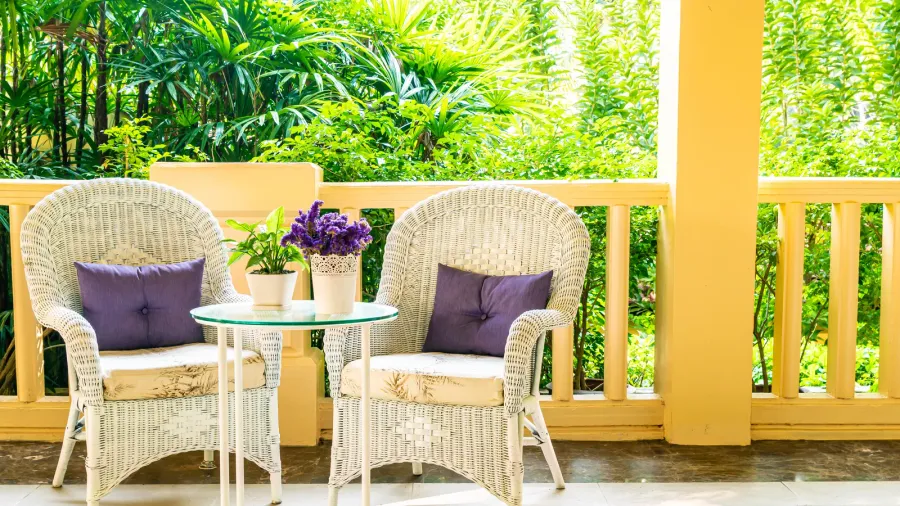 Two white rattan chairs with cushions and a small table with potted plant and flowers on a patio.