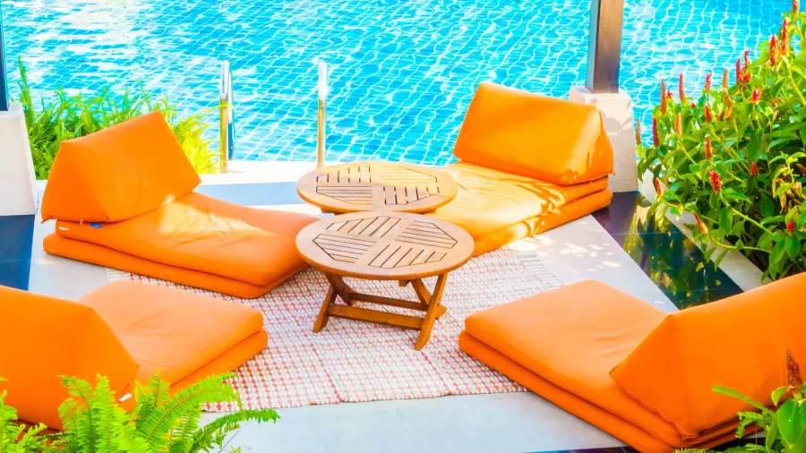 Orange lounge cushions arranged around a wooden table near a sparkling pool.