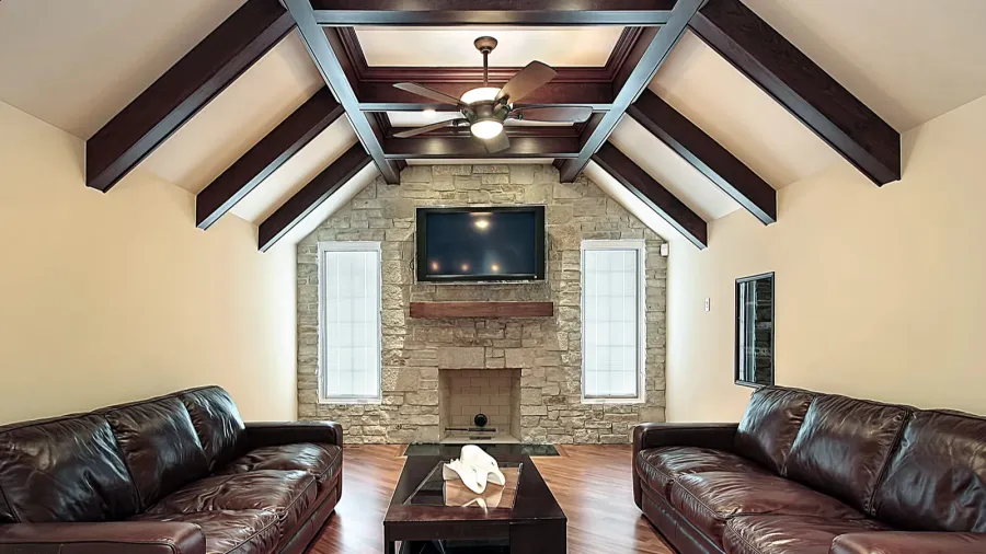 A modern living room with a vaulted ceiling, wooden beams, a stone fireplace, and black leather sofas facing a wall-mounted TV.