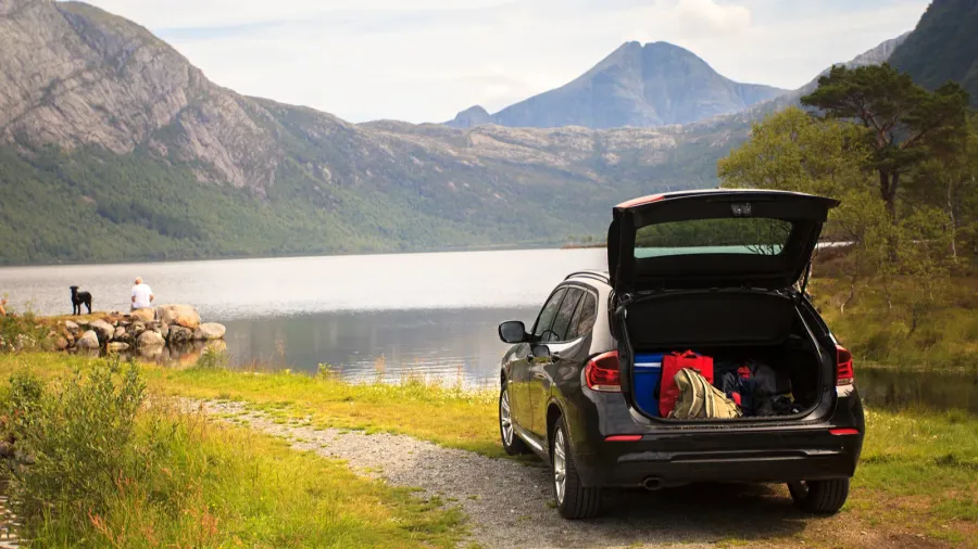 A black SUV parked near a lake with mountains in the background, open trunk showing gear inside.
