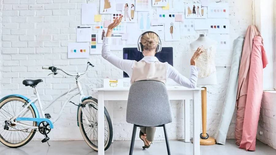 A woman sitting at a desk with a computer, wearing headphones, raising both arms, surrounded by fashion sketches and fabric samples.