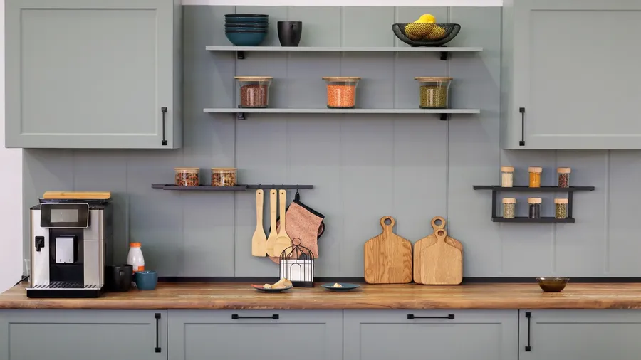 A stylish kitchen with gray cabinets, coffee maker, cutting board, and floating shelves with spices and bowls.