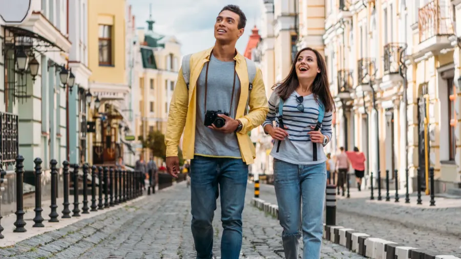 Two travelers walk cobblestone street lined with ornate colorful buildings, balconies, street lamps, and spring atmosphere.