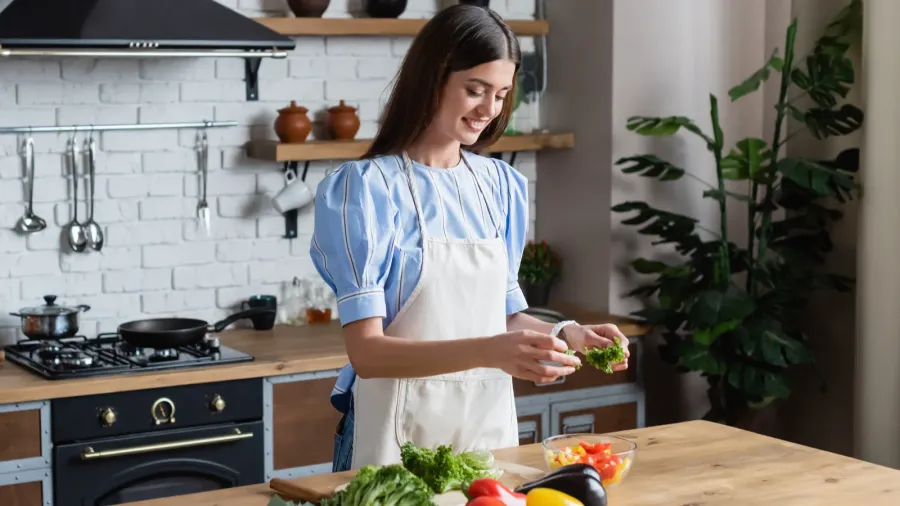 happy young adult woman in apron preparing vegetables salad in