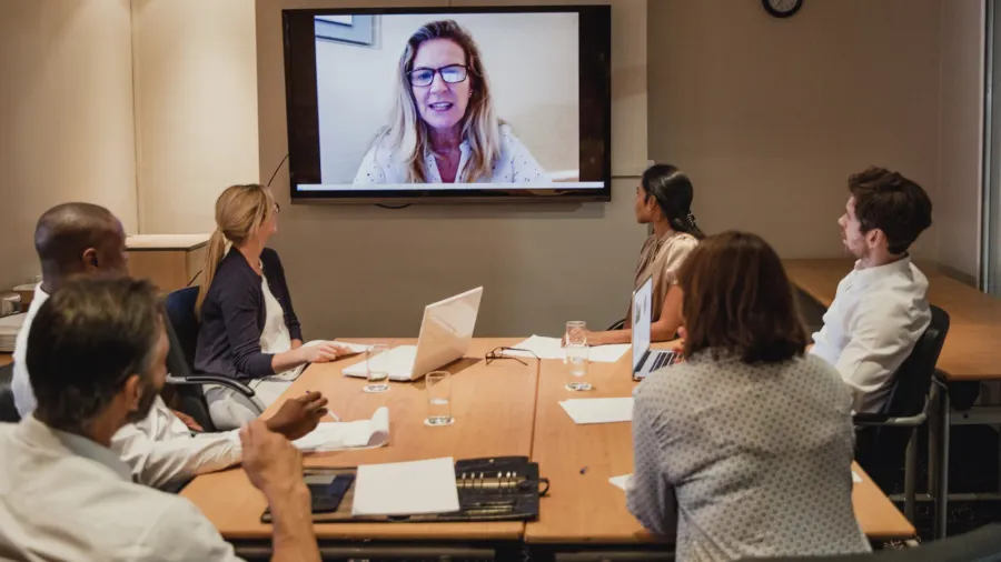 A group of people is seated at a table in a conference room, participating in a video call. One person is visible on a screen, and several others are engaged, with laptops and papers on the table.