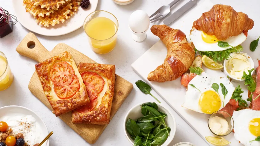 A white table with different brunch items such as toast, eggs, waffles, juice, and croissant.