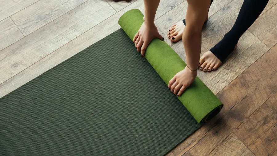 A person rolling up a green yoga mat on a wooden floor, with bare feet visible.