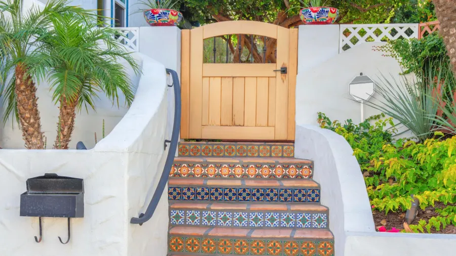 A wooden gate at the top of an outdoor staircase with colorful patterned tile risers, flanked by white walls, green plants, and a mailbox.