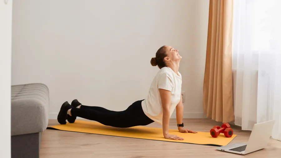 Indoor shot of fit woman making cobra pose on yoga mat, lying on her belly, places her palms under the shoulders, helping herself with hands lift the back up, wearing white T-shirt and black leggins.