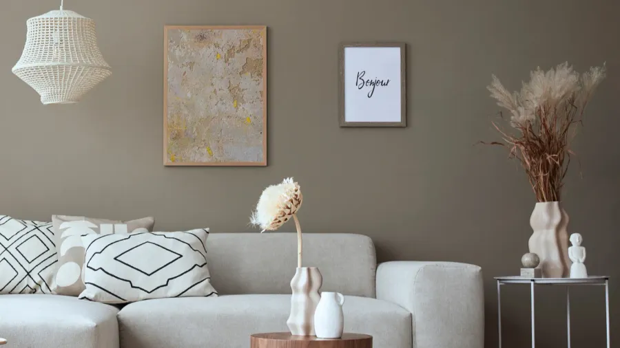 A stylish, neutral living room interior with abstract artwork and framed poster on a soft gray wall, white sofa with throw pillows, and wooden coffee table and side table with sculpted decor accents.