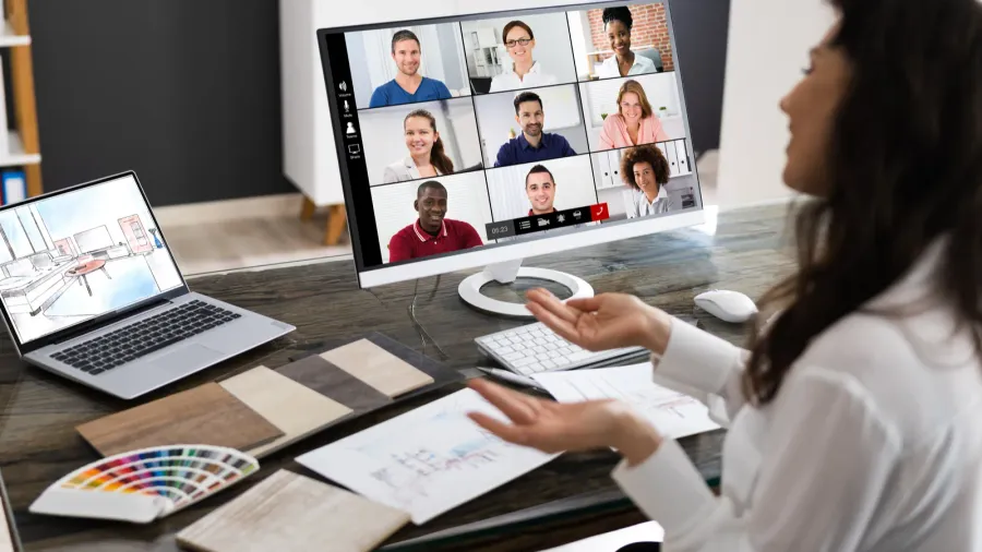 Interior designer at a desk on a video call with a team, visible on a large monitor in a grid layout. The desk holds floor plans, color swatches, wood samples, and an open laptop displaying a 3D room layout.