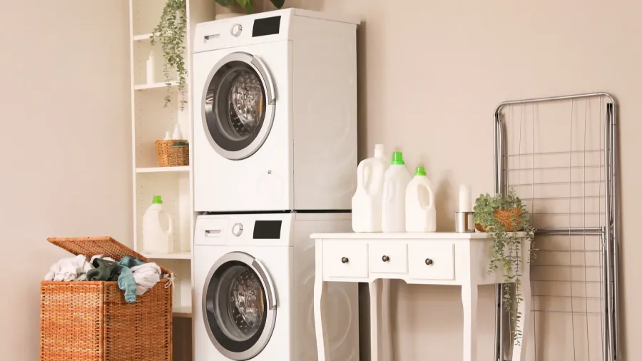 A laundry room with washing machine and dryer, a white table with detergents and cleaning supplies, a shelf with plant and storage baskets, a clothes hanging rack, and a woven laundry basket.