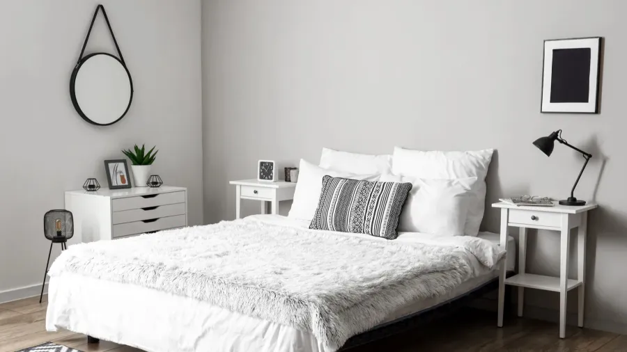 A white bedroom interior with cozy bed, nightstands, round hanging mirror, and white shelf with drawers and decor accents on it.