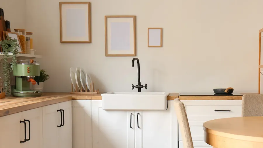 A right kitchen interior with white cabinetry, butcher block countertop, framed posters, coffee machine, and farmhouse sink with matte black tap.