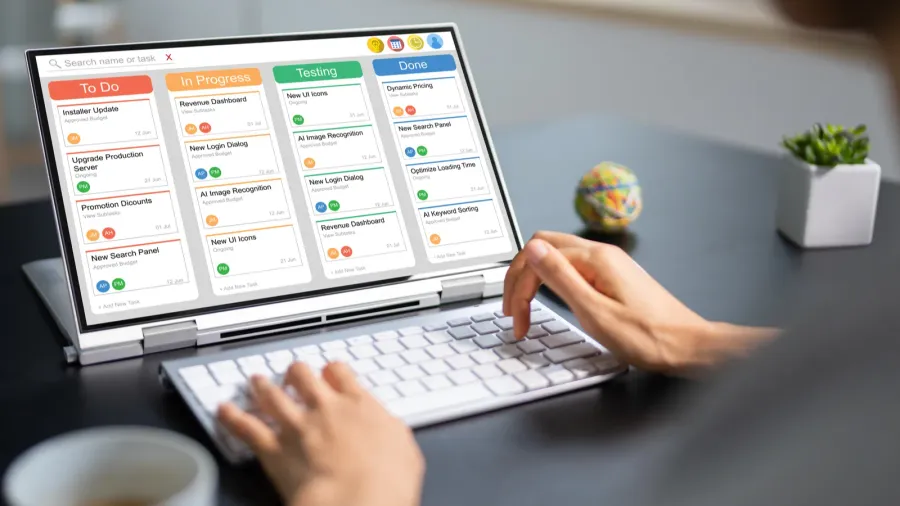 A person using a laptop displaying a colorful task board interface with columns labeled "To Do," "In Progress," "Testing," and "Done" representing an automated project management tool for organizing design tasks and streamlining workflow. A coffee cup and small plant sit nearby on the desk.