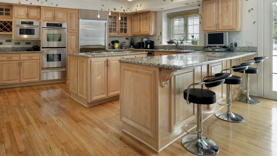 A bright kitchen interior with honey-toned oak wood cabinetry, bar stools, and granite countertop.