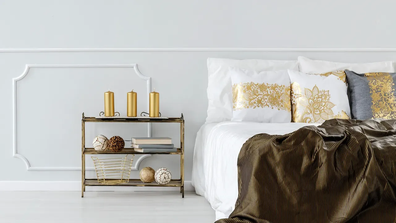 A bedroom interior with a bed with white bedsheet and white and gray pillows with gold imrprint, and a side table with golden candles and decor accents.