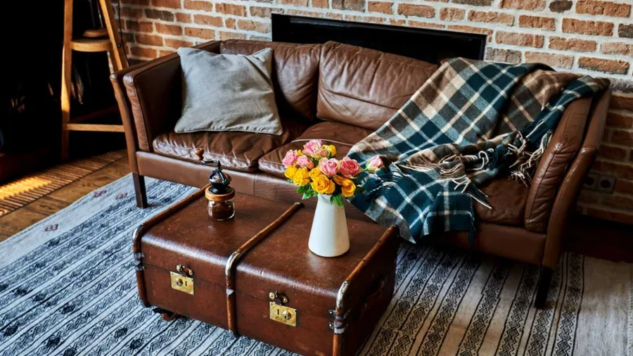 A cozy living room with a brown leather sofa, a plaid blanket, a grey pillow, a wooden trunk coffee table, and a vase of colorful flowers.