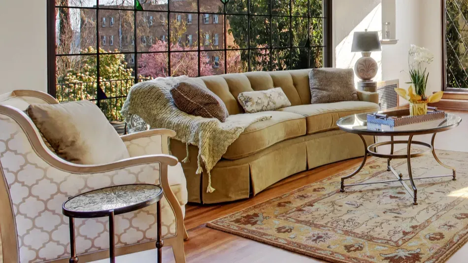 A sunlit living room with a curved sofa, patterned rug, and large bay window.