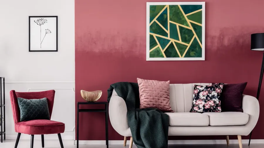 A stylish living room featuring a beige sofa with a dark green throw and pink pillow, a red velvet armchair, a black side table with a gold bowl, and a striking ombre wall transitioning from white to burgundy, adorned with geometric and botanical art.