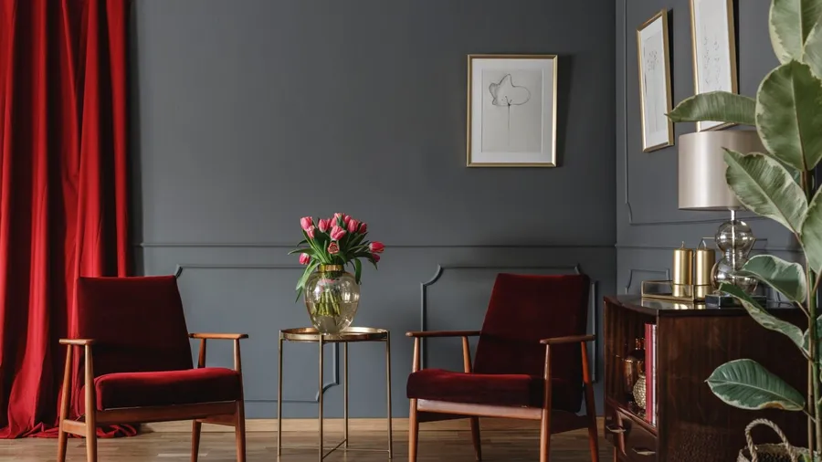 A living room with two dark red velvet armchairs with a small gold side table, pink tulips, and framed artworks on dark gray walls.