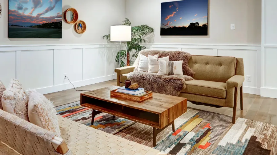 A living room interior with sofa with faux fir throw blanket and cushions, wooden coffee table, artwork on the wall, floor lamp, and rug.