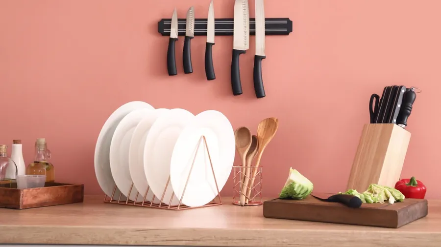A kitchen counter with plates rack, cutting board, wooden spoons in a holder, and a magnetic knife strip on pink wall.