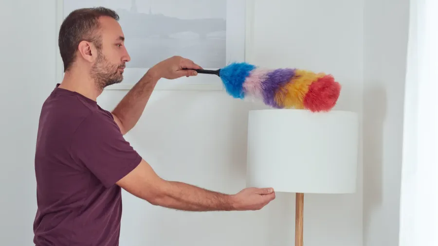 A man dusting a lamp with a colorful duster in a bright room.