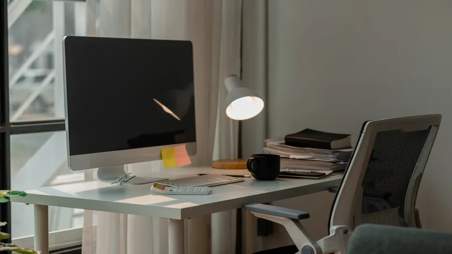 A home office with a table with computer, cup, books, and a table lamp, and a chair.
