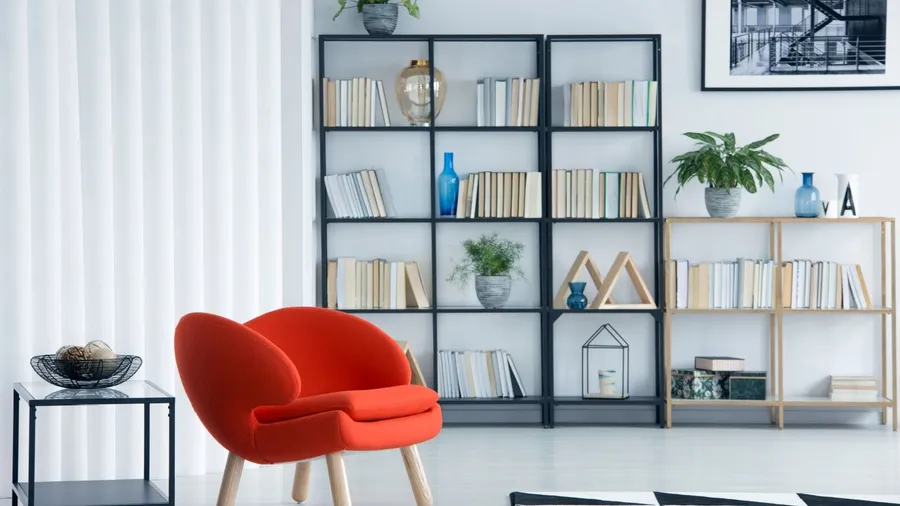 A living room interior with red armchair and metallic and wooden bookshelves with books, vases, potted plants, and decor accents.