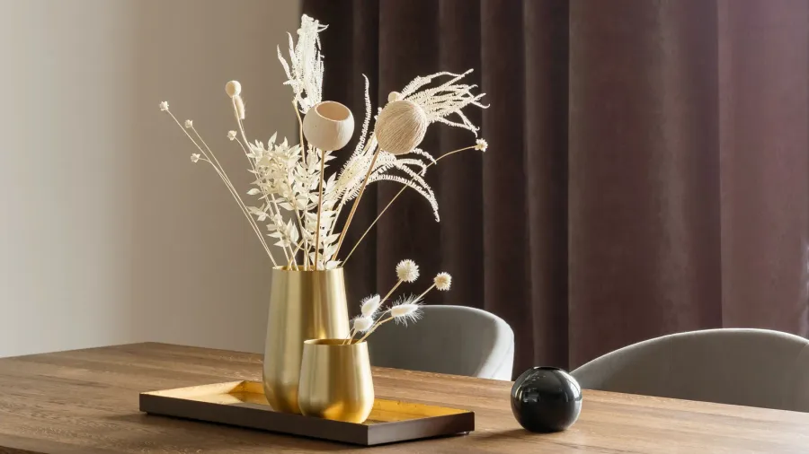 Metallic gold vases on a tray with faux branches placed on a wooden dining table.