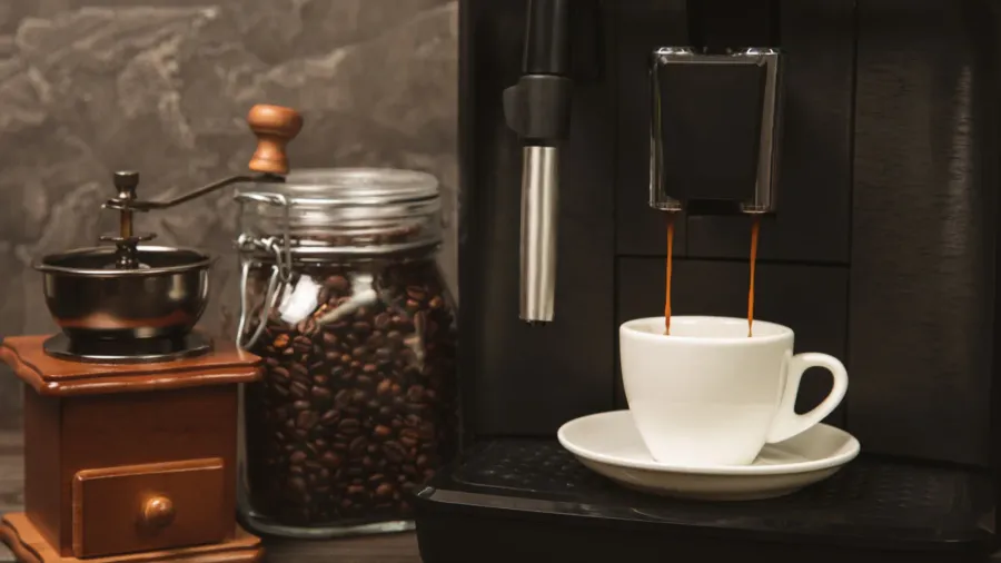 A white coffee machine with a glass jar of coffee beans, a manual coffee grinder, and a cup of freshly brewed coffee.