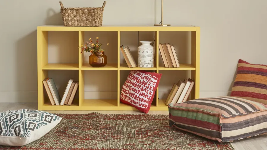 A small bookshelf with books, flowers in a vase, cushion, woven basket, and ceramic jar.