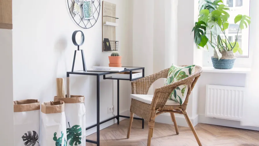 A home office interior with glass-top desk, woven chair with cushion, mesh grid on the wall, and storage baskets next to it.