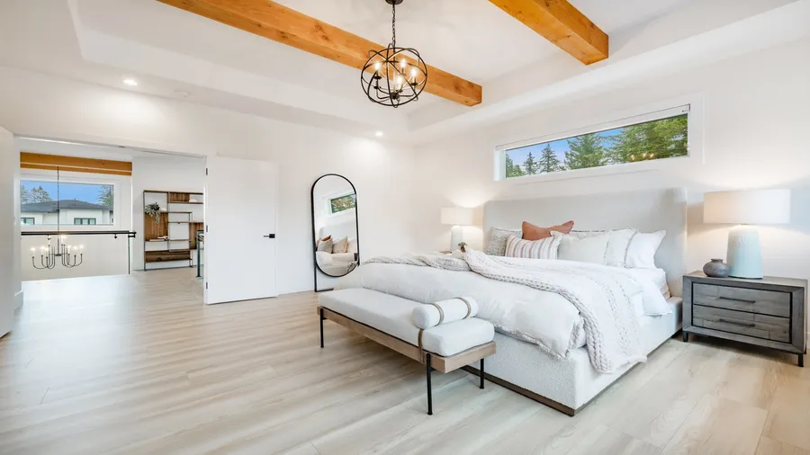 A spacious white bedroom interior with cozy bed with white bedding, bed bench, side tables, mirror, and exposed wood beams ceiling.